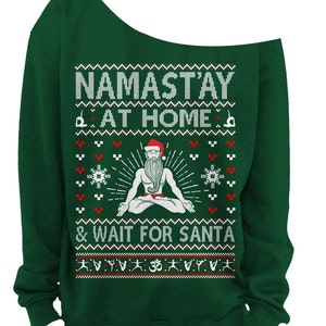 May include: A dark green off-the-shoulder sweatshirt with a festive design. The design includes the text "NAMAST'AY AT HOME & WAIT FOR SANTA" with a meditating Santa Claus figure and snowflake and yoga pose graphics.