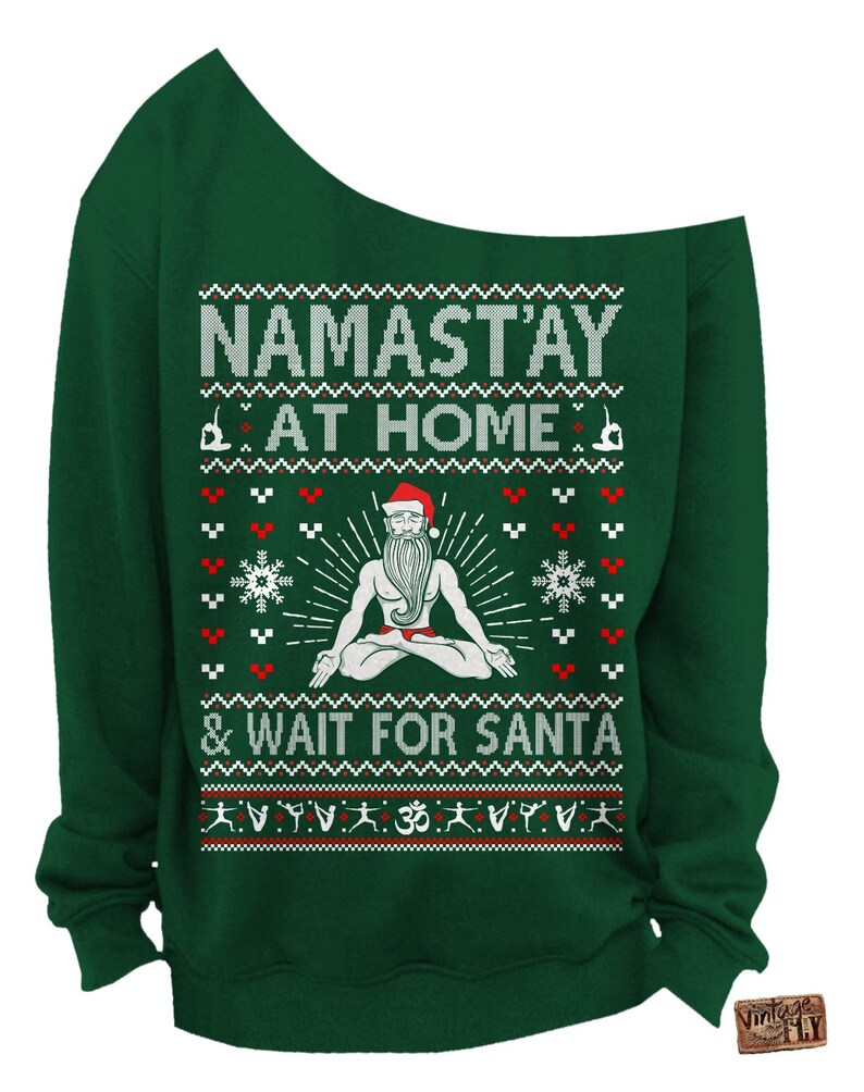 May include: A dark green off-the-shoulder sweatshirt with a festive design. The design includes the text "NAMAST'AY AT HOME & WAIT FOR SANTA" with a meditating Santa Claus figure and snowflake and yoga pose graphics.