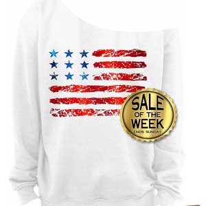 May include: White off-the-shoulder sweatshirt featuring a distressed American flag design in red and blue. The flag has blue stars and red stripes. A gold seal with the text "SALE OF THE WEEK ENDS SUNDAY" is on the right side.