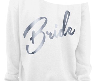 May include: A white off-the-shoulder sweatshirt featuring the word "Bride" in a metallic silver script. The top has long sleeves and a relaxed fit, suitable for bridal wear or a hen party.
