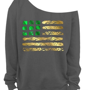 May include: Dark gray off-the-shoulder sweatshirt featuring a gold and green design. The design resembles a flag with gold stripes and green shamrocks. The sweatshirt has a relaxed fit and a unique neckline.