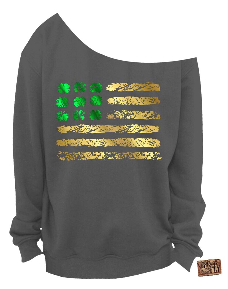May include: Dark gray off-the-shoulder sweatshirt featuring a gold and green design. The design resembles a flag with gold stripes and green shamrocks. The sweatshirt has a relaxed fit and a unique neckline.