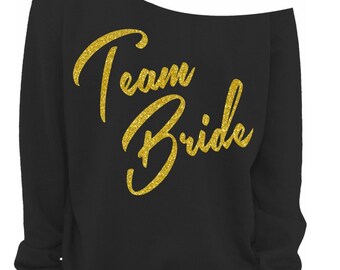 May include: Black off-the-shoulder sweatshirt with gold glitter text reading "Team Bride". The long-sleeved sweatshirt has a relaxed fit, ideal for a bridal party. The text is in a cursive font.