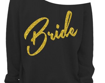 May include: Black off-the-shoulder sweatshirt with the word "Bride" in gold glitter script across the front. The sweatshirt has long sleeves and a relaxed fit. A small "Vintage Fly" logo is in the bottom right corner.