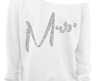 May include: White off-the-shoulder sweatshirt with the word "Mrs." in silver glitter. The sweatshirt has long sleeves and a relaxed fit. A small brown tag with the words "Vintage Fly" is in the bottom right corner.