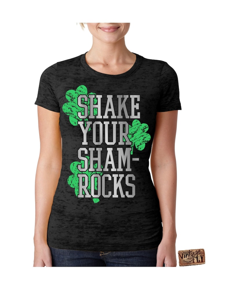 May include: Black t-shirt with the text "SHAKE YOUR SHAMROCKS" in white and green. The design includes two green shamrock graphics. The shirt is a crew neck style.