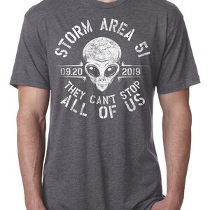 May include: Dark gray t-shirt with a white graphic featuring an alien head and the text "STORM AREA 51 09.20 2019 THEY CAN'T STOP ALL OF US". The shirt has short sleeves and a crew neck.