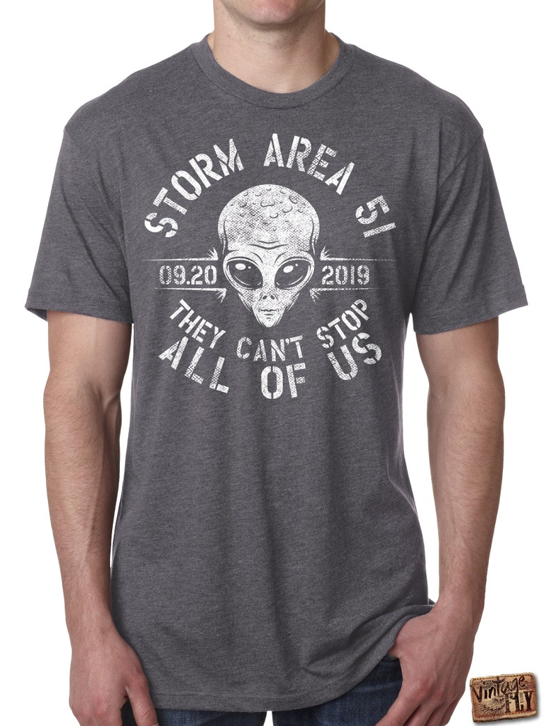 May include: Dark gray t-shirt with a white graphic featuring an alien head and the text "STORM AREA 51 09.20 2019 THEY CAN'T STOP ALL OF US". The shirt has short sleeves and a crew neck.