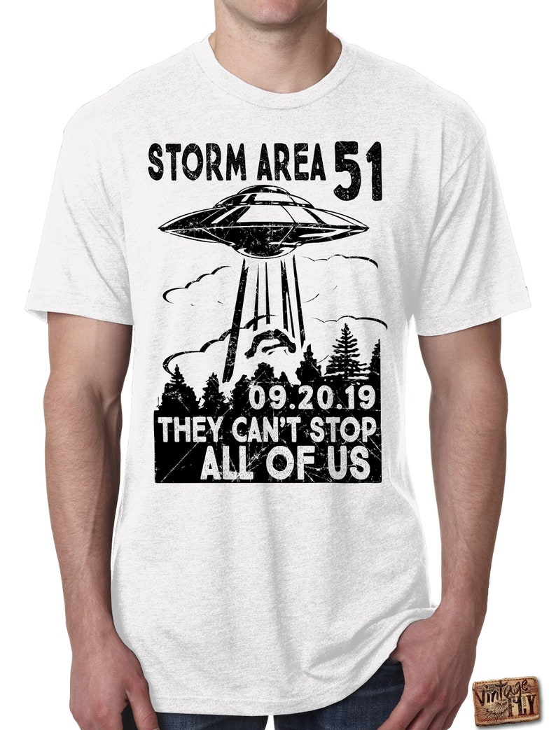 May include: White t-shirt with a black graphic design. The design features a UFO above a forest silhouette, with the text "STORM AREA 51" and "09.20.19 THEY CAN'T STOP ALL OF US". The shirt is a crew neck style.