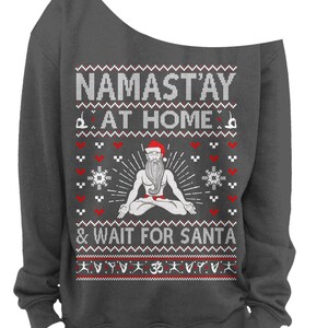 May include: Dark gray off-the-shoulder sweatshirt with a festive design. The text reads "NAMAST'AY AT HOME & WAIT FOR SANTA" above a graphic of Santa in a yoga pose. The design includes red hearts, snowflakes, and yoga poses.