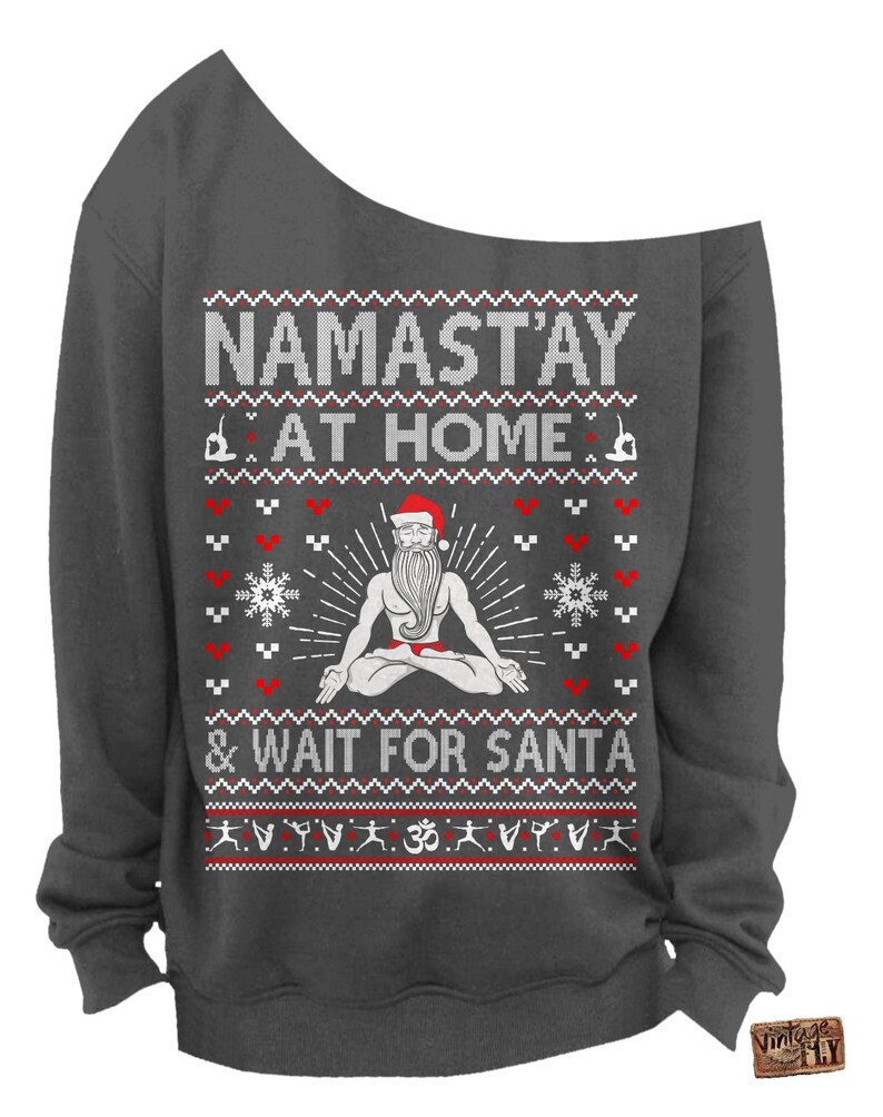 May include: Dark gray off-the-shoulder sweatshirt with a festive design. The text reads "NAMAST'AY AT HOME & WAIT FOR SANTA" above a graphic of Santa in a yoga pose. The design includes red hearts, snowflakes, and yoga poses.