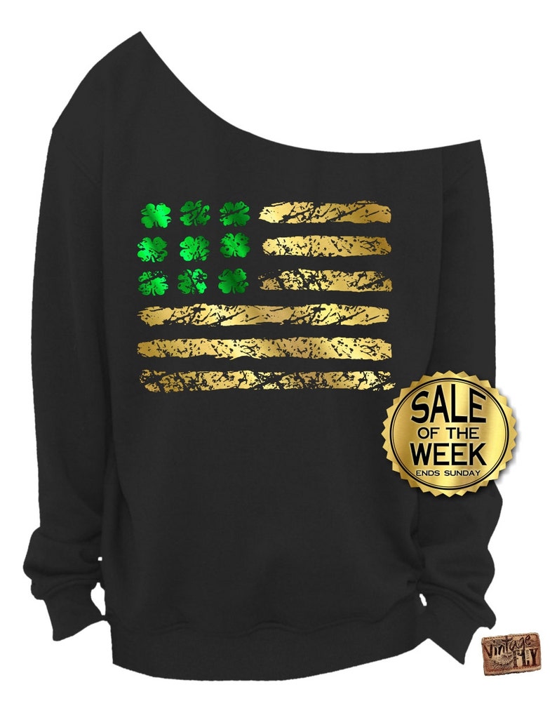 May include: Black off-the-shoulder sweatshirt with a gold and green graphic design. The design resembles a flag with gold stripes and green shamrocks. A gold sticker on the right side says "SALE OF THE WEEK ENDS SUNDAY."