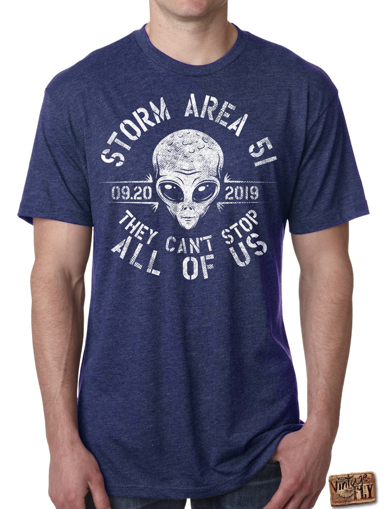 May include: Navy blue t-shirt with a white graphic of an alien head and the text "STORM AREA 51 09.20 2019 THEY CAN'T STOP ALL OF US". The shirt has short sleeves and a crew neck.