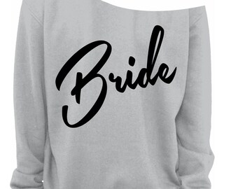 May include: A pale grey off-the-shoulder sweatshirt featuring the word "Bride" in large, black cursive script. The garment has long sleeves and a relaxed fit, suitable for a bridal celebration.
