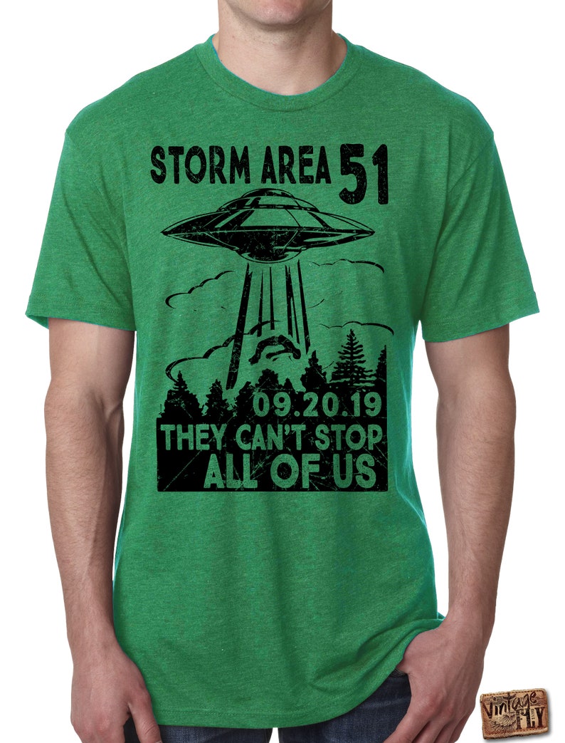 May include: Green t-shirt with a black graphic of a UFO above a forest, with the text "STORM AREA 51" and "09.20.19 THEY CAN'T STOP ALL OF US". The shirt has short sleeves and a crew neck.