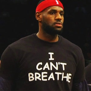 May include: A person wearing a black t-shirt with the words "I CAN'T BREATHE" in white block letters. The person is also wearing a red headband with a white logo.