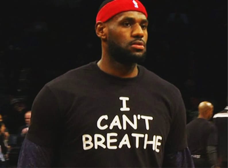May include: A person wearing a black t-shirt with the words "I CAN'T BREATHE" in white block letters. The person is also wearing a red headband with a white logo.