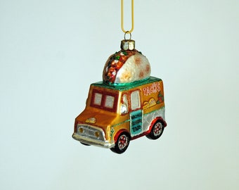 May include: A festive glass ornament shaped like a taco truck. The truck is gold and silver with red wheels and the word "TACOS" on the side. A detailed taco sits on top. The ornament is suspended by a gold string.