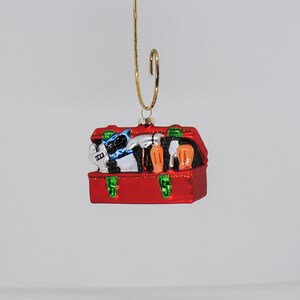 May include: A red Christmas ornament shaped like a suitcase, with green clasps and a gold hanging hook. The suitcase is filled with miniature items, including a black and white cat, and orange bottles. The ornament is suspended against a white background.