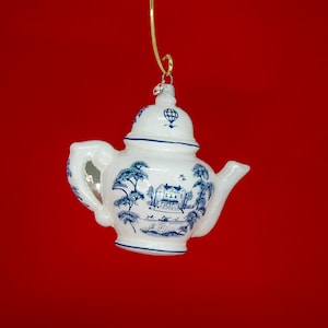 May include: White ceramic teapot Christmas ornament with blue landscape design. The ornament features a detailed illustration of trees, a house, and figures, with blue trim. A gold-colored hook is attached for hanging.