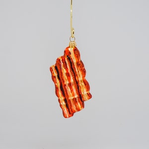 May include: A bacon-shaped Christmas ornament. The ornament is a reddish-orange color with gold accents, resembling cooked bacon. It hangs from a gold-colored metal loop.