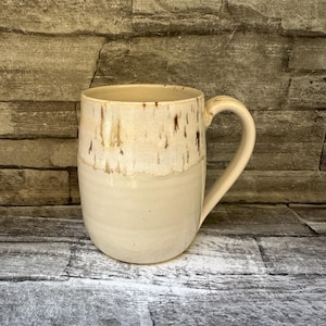 May include: A ceramic mug with a smooth, cream-colored base and a textured, speckled upper section featuring brown drip-like glaze patterns. The mug has a rounded body and a curved handle on the right side, showcasing a two-tone finish.