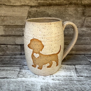 May include: A ceramic mug with a speckled cream finish and a large handle. The front features a tan-colored silhouette of a dachshund dog with a small white heart shape on its chest. The mug has a rounded body and a smooth, glazed texture.