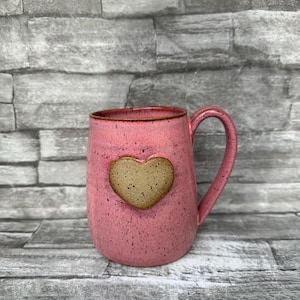 May include: Pink ceramic mug with a brown heart-shaped accent. The mug has a speckled texture and a brown rim. The handle is curved and matches the mug's color. A charming and rustic piece for enjoying your favorite beverage.