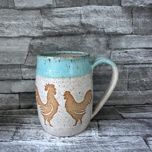 May include: A ceramic coffee mug with a light blue rim and handle, and a speckled white body. Two brown rooster silhouettes are featured on the mug's front. The mug is designed for hot beverages.