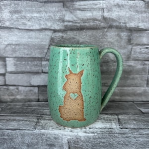 May include: A speckled turquoise ceramic mug with a brown silhouette of a dog and a heart. The mug has a curved handle and is set against a gray brick background. The mug is a handmade item.