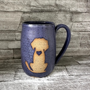 May include: A blue ceramic mug with a tan dog silhouette and heart detail. The mug has a curved handle and a speckled finish. The dog illustration is centered on the mug's body.