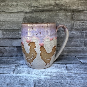 May include: A ceramic mug with a speckled cream base and a lavender drip glaze at the rim. The mug features two brown rooster silhouettes facing each other on the front. It has a sturdy handle on the right side and a smooth, rounded body shape.