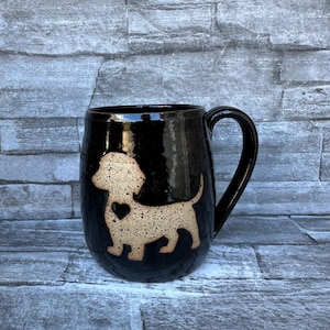 May include: Black ceramic mug with a tan dachshund dog design and a heart. The mug has a curved handle and a glossy finish. The dog design is centered on the mug.