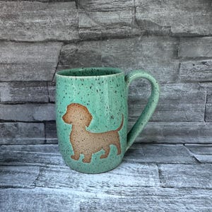 May include: A ceramic mug with a speckled mint green glaze featuring a tan-colored silhouette of a dachshund dog on the front. The mug has a curved handle on the right side and a smooth, rounded body shape with a glossy finish throughout the exterior.