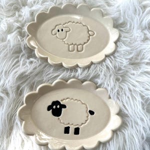 May include: Two white ceramic oval-shaped dishes with scalloped edges. Each dish features a raised sheep design. One sheep is white with black eyes and the other sheep is black with white eyes.