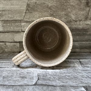 Pode incluir: Uma caneca de cer&acirc;mica com acabamento bege salpicado e textura canelada no exterior e na pega. O interior &eacute; liso e de cor clara, apresentando uma base circular e estrias verticais ao longo das paredes internas do recipiente.