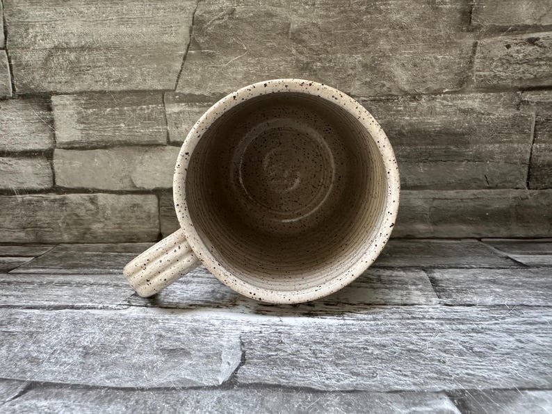 Pode incluir: Uma caneca de cer&acirc;mica com acabamento bege salpicado e textura canelada no exterior e na pega. O interior &eacute; liso e de cor clara, apresentando uma base circular e estrias verticais ao longo das paredes internas do recipiente.