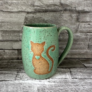 May include: A handmade ceramic mug with a speckled turquoise glaze. The mug features a brown cat silhouette with a heart detail. The handle and interior match the exterior. This unique mug is perfect for coffee, tea, or any beverage.
