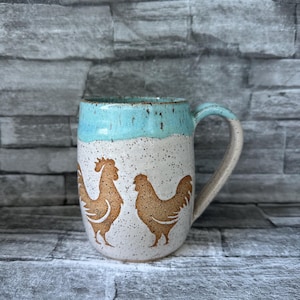 May include: A ceramic mug with a light blue top and a speckled white base. Two brown rooster silhouettes are featured on the front. The handle is a light beige color. The mug is designed for hot beverages.