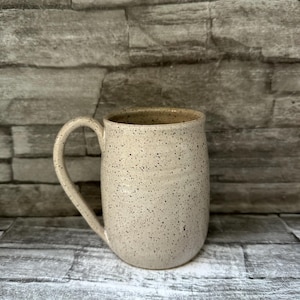 Pode incluir: Uma caneca de cer&acirc;mica com um esmalte de cor creme salpicado e um corpo liso e arredondado. A caneca apresenta uma &uacute;nica pega curva no lado esquerdo e uma abertura larga. A superf&iacute;cie exterior est&aacute; coberta com pequenas manchas castanhas.