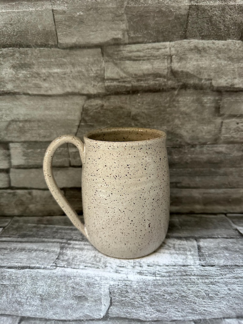 Pode incluir: Uma caneca de cer&acirc;mica com um esmalte de cor creme salpicado e um corpo liso e arredondado. A caneca apresenta uma &uacute;nica pega curva no lado esquerdo e uma abertura larga. A superf&iacute;cie exterior est&aacute; coberta com pequenas manchas castanhas.