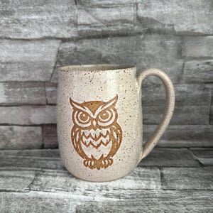 May include: A speckled ceramic mug with a brown owl design. The mug has a large handle and a beige background with dark brown speckles. The owl is centered on the mug and has detailed features.