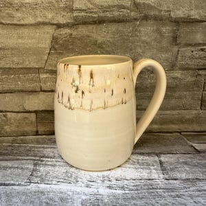 May include: A ceramic mug with a smooth, cream-colored base and a textured, dripped brown glaze around the upper rim. The mug features a rounded body and a large, curved handle on the right side, showcasing a handcrafted aesthetic with subtle variations.