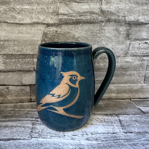 May include: A handmade ceramic mug with a blue glaze and a bird design. The mug has a dark green handle and a cylindrical shape. The bird is a light brown colour and is centered on the mug.