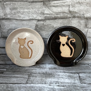 May include: Two ceramic spoon rests shaped like cats. One is white with brown speckles and a brown cat with a heart on its chest. The other is black with a white cat with a heart on its chest.