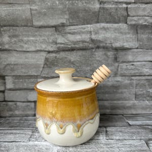May include: A ceramic honey pot with a lid and a wooden honey dipper. The pot is glazed in shades of yellow, brown, and white, with a wavy design at the bottom. The lid is off-white with a small knob.