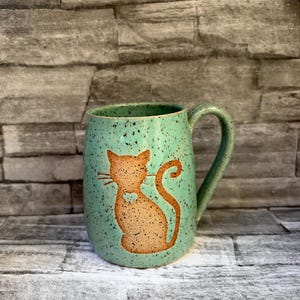 May include: A speckled mint green ceramic mug featuring a tan silhouette of a sitting cat with a small heart-shaped cutout on its chest. The mug has a curved handle on the right side and a smooth, rounded body with dark brown speckles throughout.