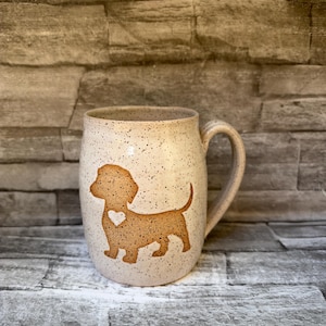 May include: A ceramic mug with a speckled cream finish featuring a brown silhouette of a dachshund dog on the front. The dog design includes a small heart-shaped cutout in the center of its body. The mug has a curved handle on the right side.