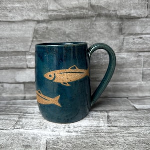 May include: A dark teal ceramic mug featuring a glossy finish and a speckled texture. The exterior is decorated with two light brown, etched fish silhouettes positioned one above the other. The mug includes a curved handle on the right side.