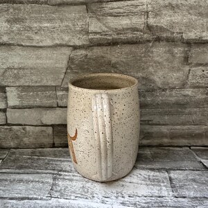 Pode incluir: Uma caneca de cer&acirc;mica bege salpicada com uma al&ccedil;a lisa e arredondada. A superf&iacute;cie externa apresenta uma cor de base creme clara com in&uacute;meras pequenas manchas castanho-escuras distribu&iacute;das por toda a pe&ccedil;a. Um gr&aacute;fico castanho &eacute; vis&iacute;vel.